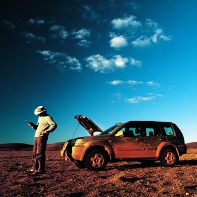 Man with a mobile phone in his hand standing next to his broken down car in the outback.